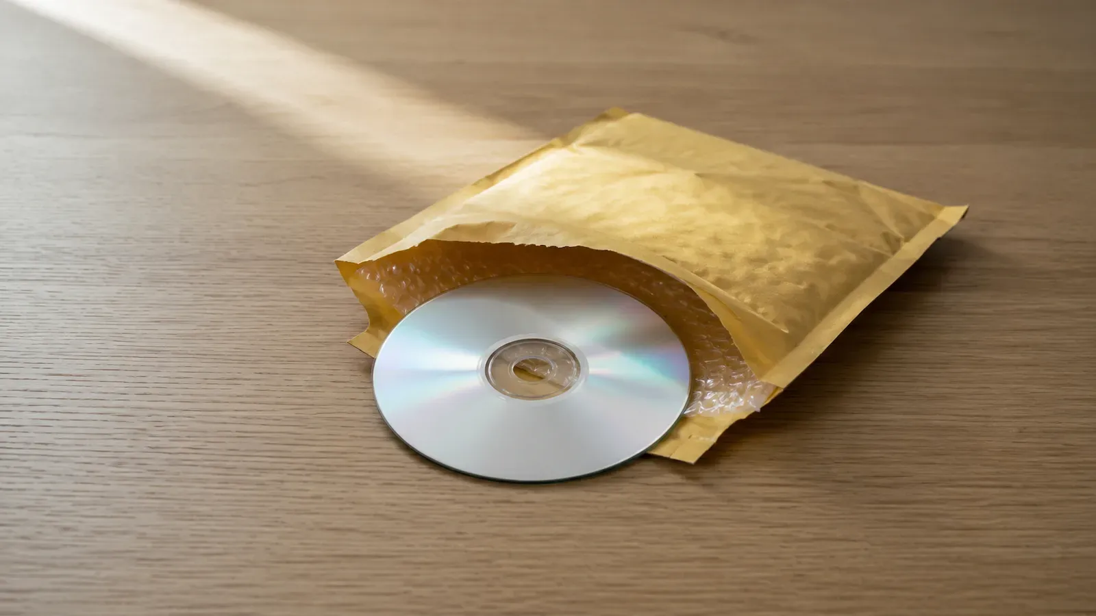 A blank recordable CD lying on top of an open padded mailing envelope on a wooden desk